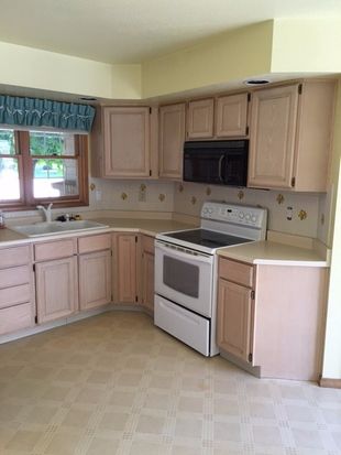 Dated kitchen with soffits, mismatched white and black appliances, lighter wood cabinets, and linoleum flooring.